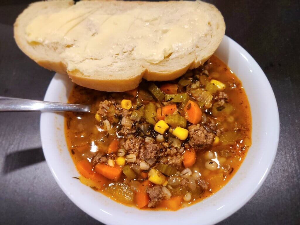 Bowl of hearty beef and barley soup with ground beef, carrots, celery, corn, and herbs, served with buttered bread on the side