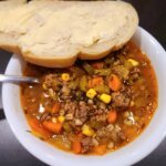 Bowl of hearty beef and barley soup with ground beef, carrots, celery, corn, and herbs, served with buttered bread on the side